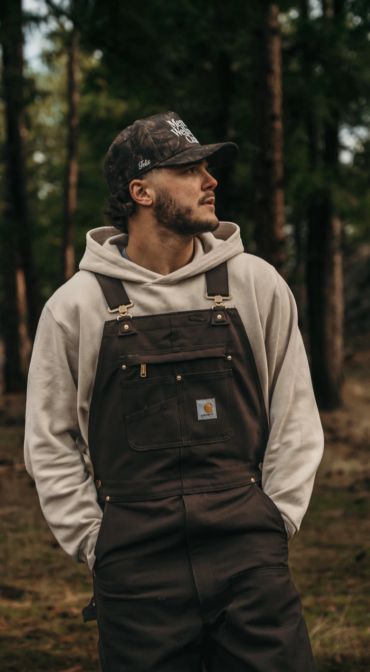 Man in brown overalls and cap in a forest.