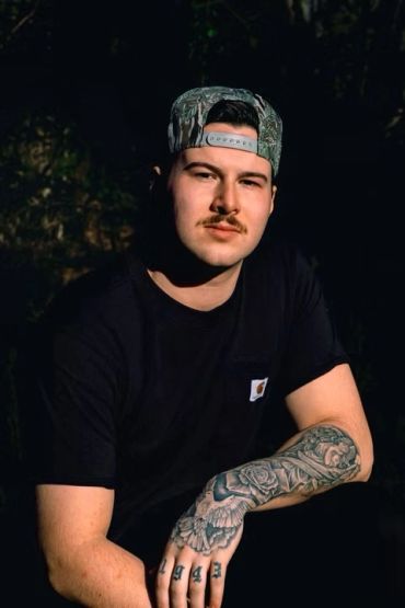 Man with a mustache and tattooed arm wearing a cap.