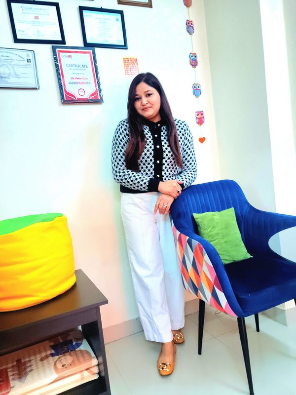 Woman standing beside a colorful chair in a cozy, certificate-decorated room.
