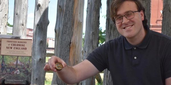 Man smiling, holding a compass in front of wooden fence.