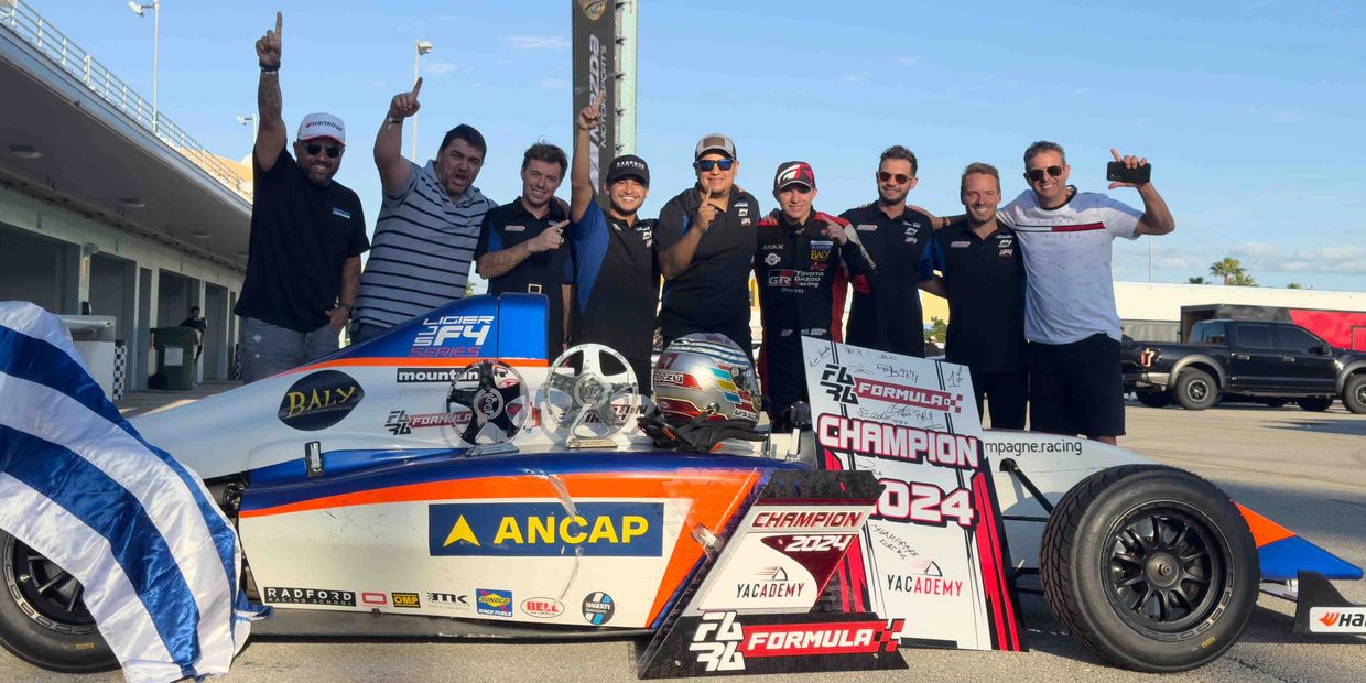 Racing team celebrates victory with their car and championship trophies in bright daylight.