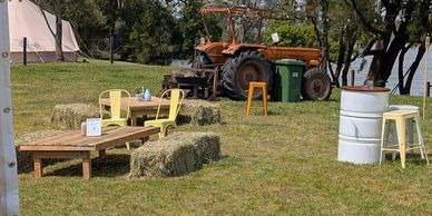 Outdoor rustic seating with hay bales, yellow chairs, and an old tractor in the background.