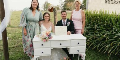 Newlywed couple holding marriage certificate with two women at outdoor wedding.