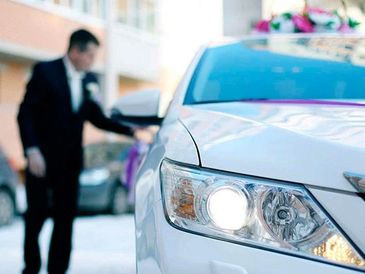 A well-dressed man opening the door of a white car with headlights on.