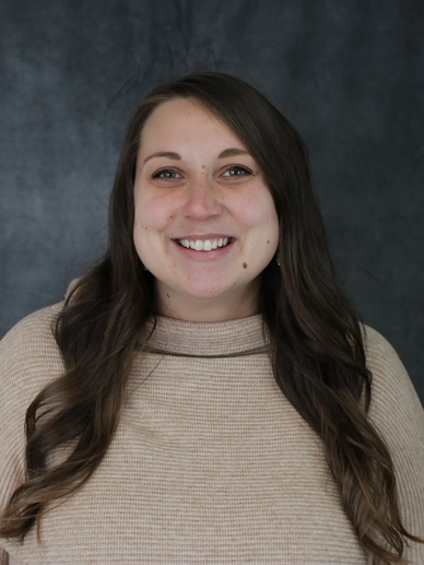 Smiling woman with long brown hair and a beige sweater against a dark background.