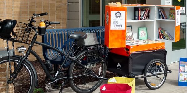 A black bike and trailer with a custom build folding bookcase full of books for people to take. 