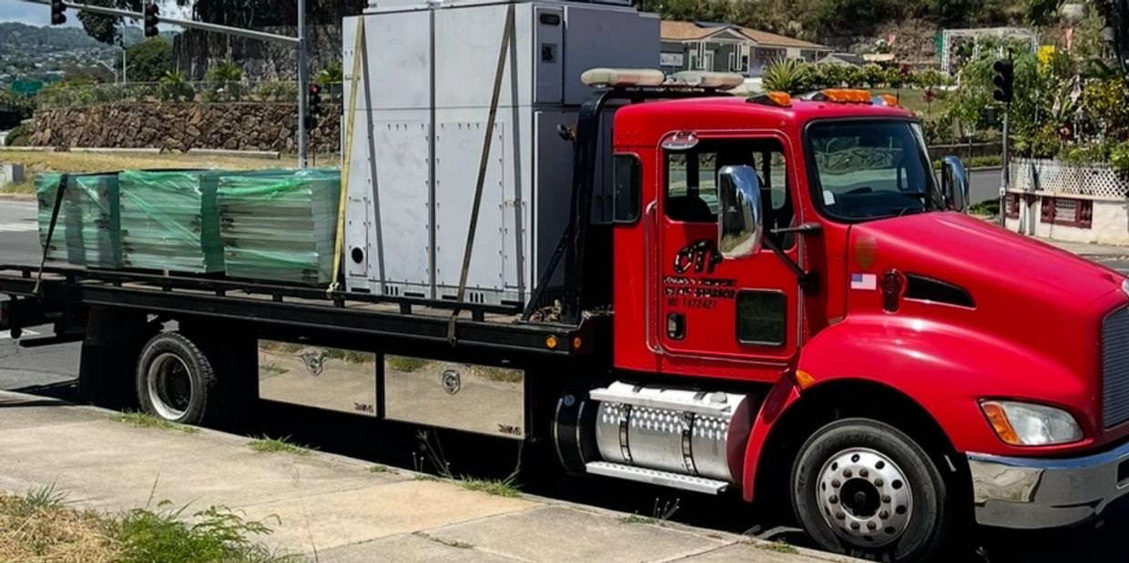 Red flatbed truck carrying large appliances and equipment on a sunny day.