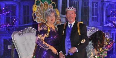 A man and woman dressed as king and queen sit on a throne at a festive event.