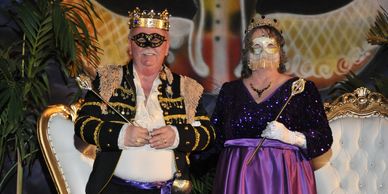 A regal couple in ornate crowns and masks sit on luxurious chairs at a themed event.