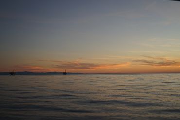 Calm ocean at sunset with sailboats on the horizon.