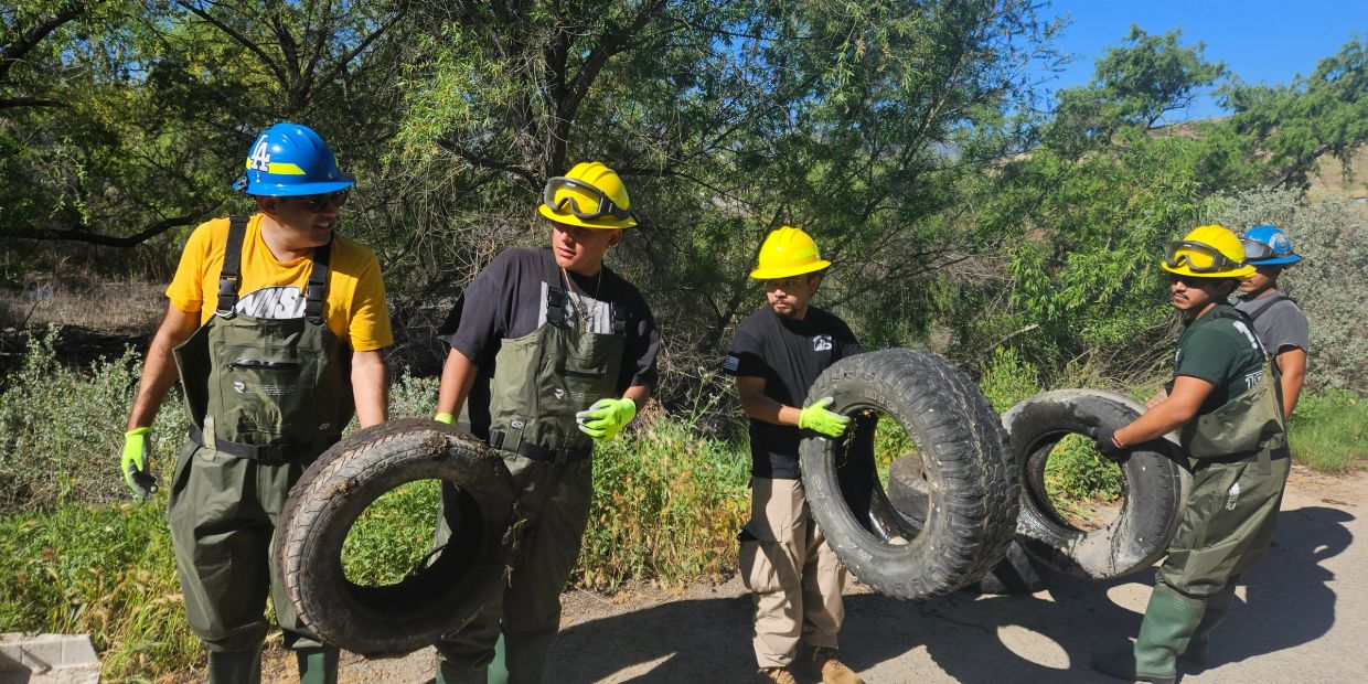 Workers wearing safety gear carry old tires in a natural outdoor setting.