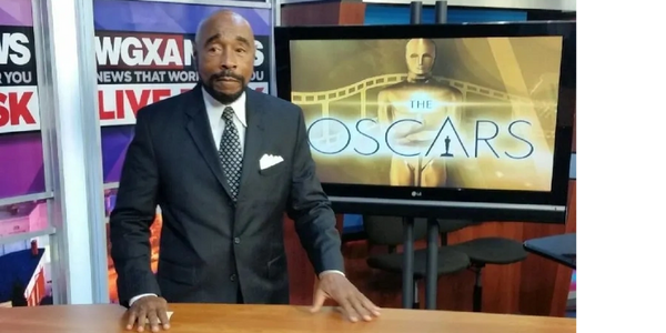 News anchor standing behind a desk in a studio with Oscars on screen.