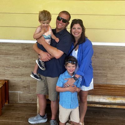 A happy family of four posing together outdoors against a yellow wall.