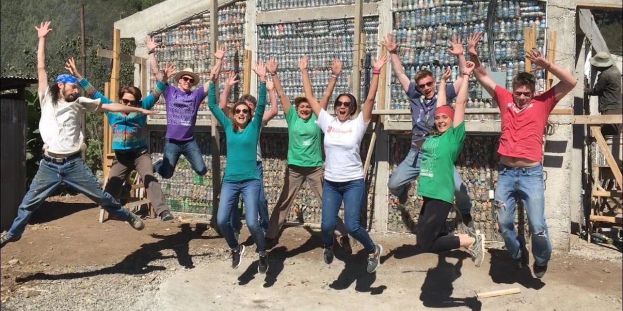 Group of people jumping joyfully outdoors in front of a wall made of plastic bottles.