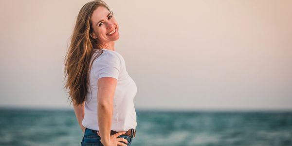Smiling woman in a white shirt and jeans stands by the sea at sunset.