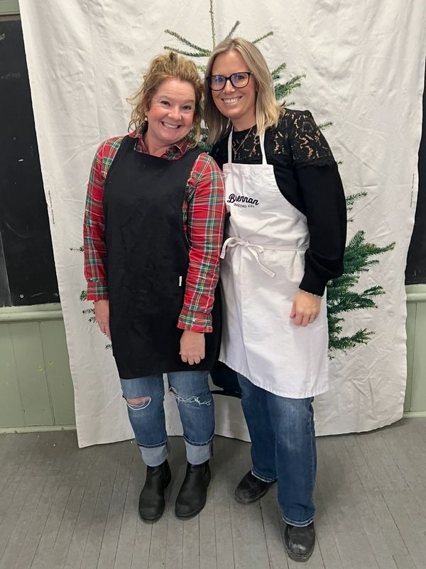 Two women wearing aprons posing cheerfully in front of a festive backdrop.