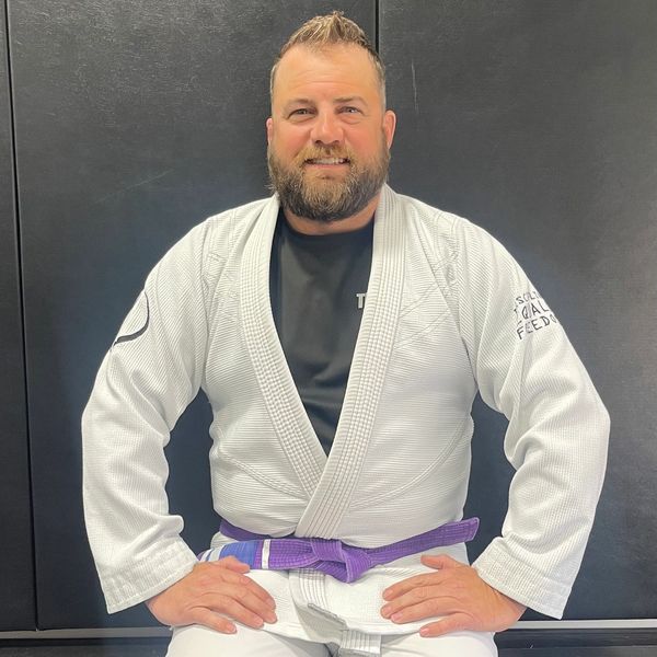 Man in a white martial arts gi with a purple belt sitting and smiling.