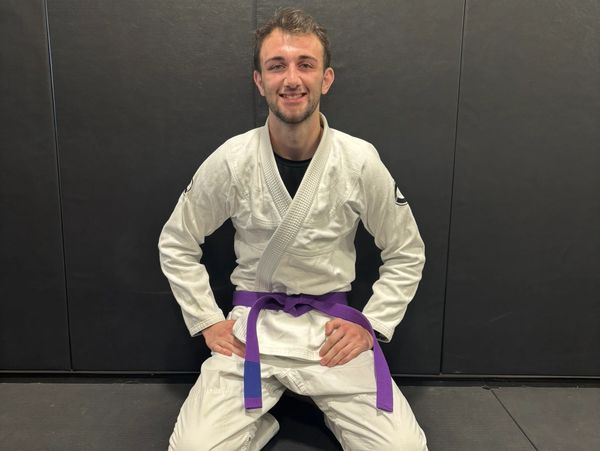 Man in a white martial arts gi with a purple belt kneeling on a mat.
