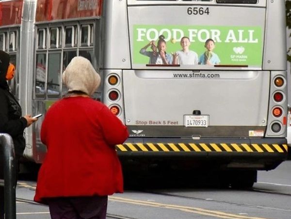 Person in red jacket walking behind a city bus with a food advertisement.