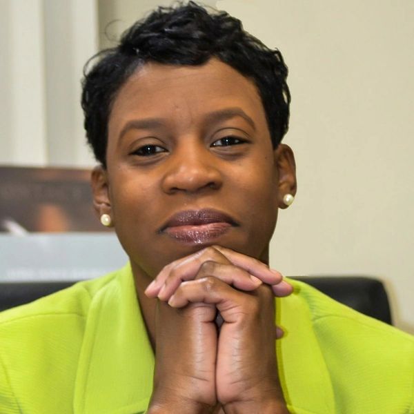 Woman in a green jacket with hands clasped, looking thoughtfully.