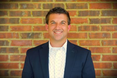 Smiling man in a navy blazer standing against a brick wall.