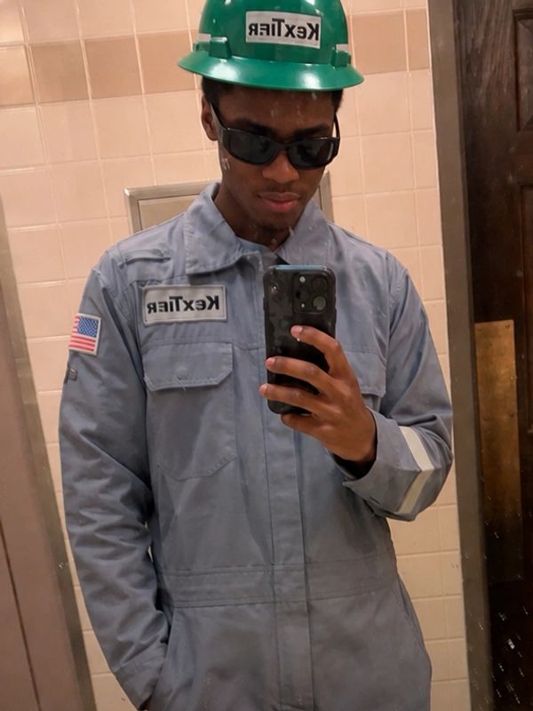 Young man in work uniform and green hard hat taking a mirror selfie.