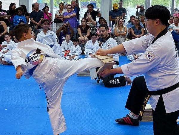 A young martial artist kicks a board held by an instructor during a class with an audience.