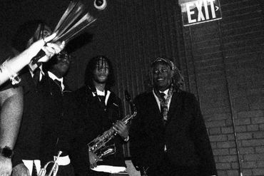A black and white photo of musicians with saxophone and trumpet under an exit sign.