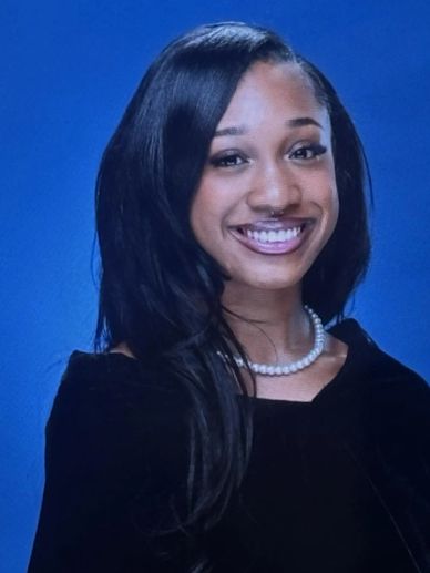 Young woman with long hair smiles wearing a pearl necklace and black top against blue backdrop.