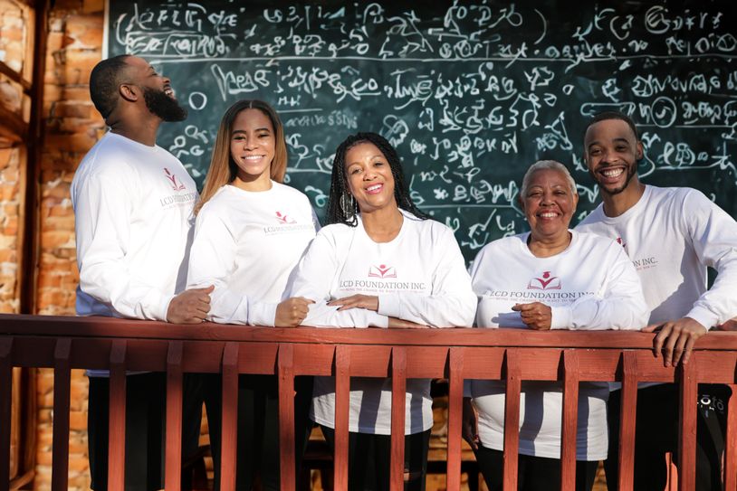 Five diverse people smiling in white foundation shirts with a chalkboard full of equations behind them.