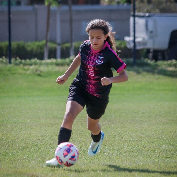 Young girl in black soccer uniform kicking a ball on a grassy field.