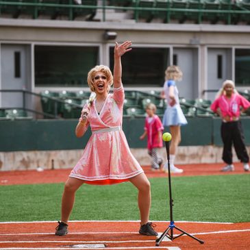 Woman in a pink dress enthusiastically pitching a ball on a baseball field.