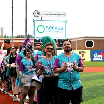 People lined up on a baseball field in colorful jerseys at a Servus Credit Union event.