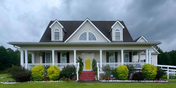 White house with yellow door and vibrant garden under cloudy sky.