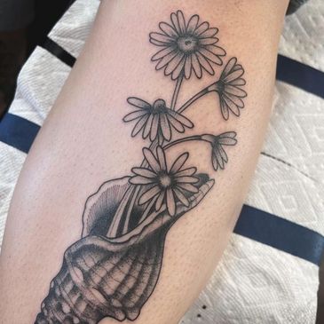 Black and white tattoo of daisies growing from a seashell on skin.