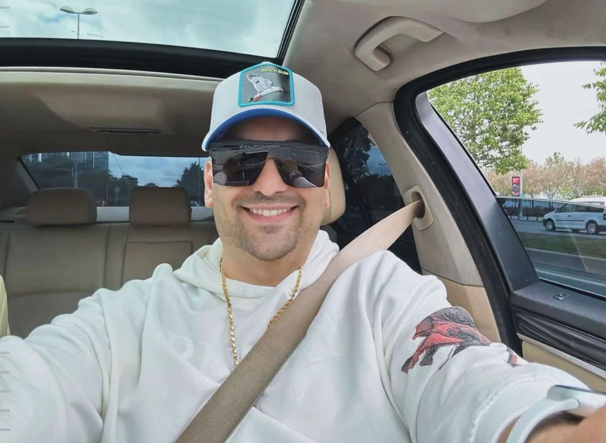 Man wearing sunglasses and a cap smiles inside a car with a sunroof.