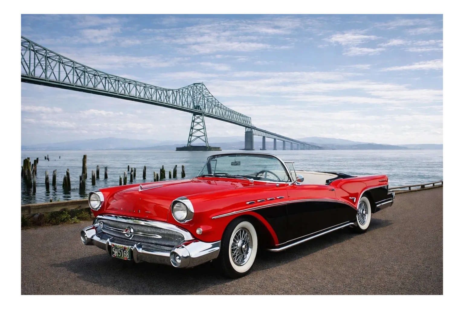 Classic red and black convertible parked near a waterfront with a bridge in the background.