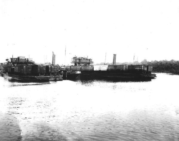 Two barges loaded with cargo containers on a river near a wooded shoreline.