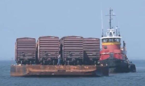 Tugboat towing a barge with four train boxcars on the water.