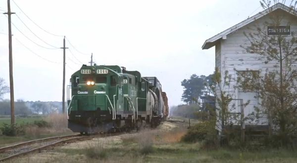 A green Canadian locomotive pulls freight cars along a rural railroad track.