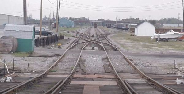 Railroad tracks diverge in an industrial area with trains and buildings nearby.