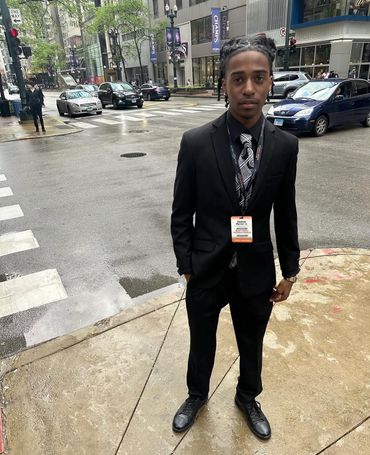 Young man in a black suit standing on a city sidewalk.