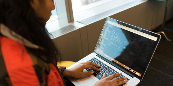 Person typing code on a laptop near a window in a cozy setting.