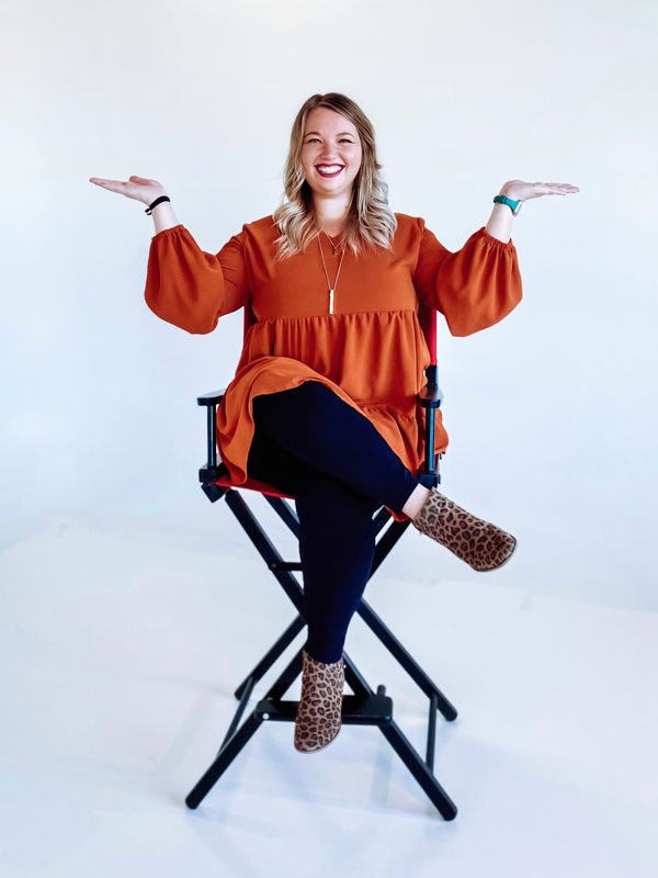 Smiling woman in an orange dress sitting on a director's chair with arms raised.