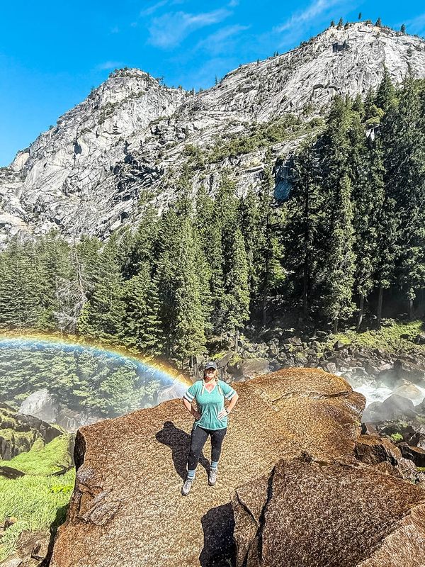 Person standing on rock with a rainbow and mountain backdrop.