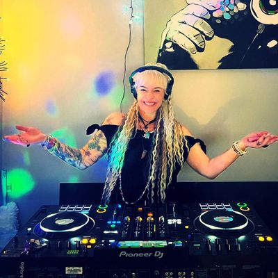 Smiling female DJ with long blonde hair playing music in a colorful room.