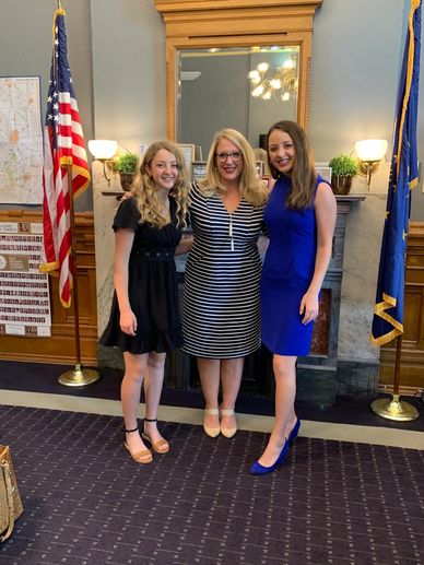 Three women smiling indoors with flags and plants in the background.