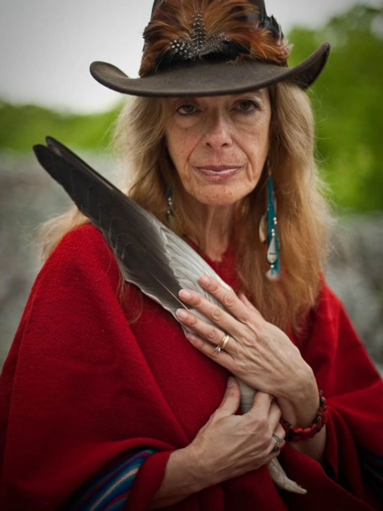 Woman in red holding large feather, wearing a hat and earrings.