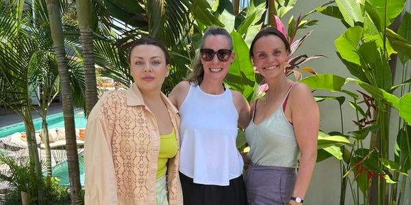 Three women standing outdoors with tropical plants in the background.