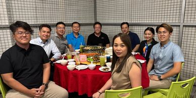 A group of nine people enjoying a meal at a round table with a red tablecloth.